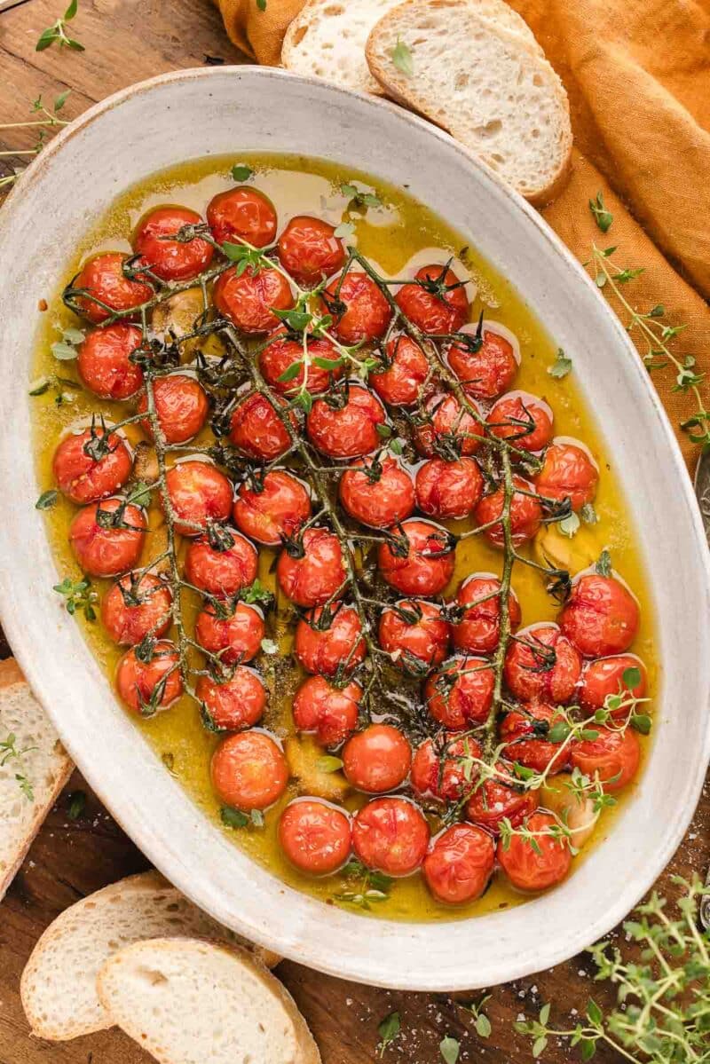 Oval dish filled with tomato confit, garnished with herbs. Slices of bread are placed around the dish on a wooden surface.