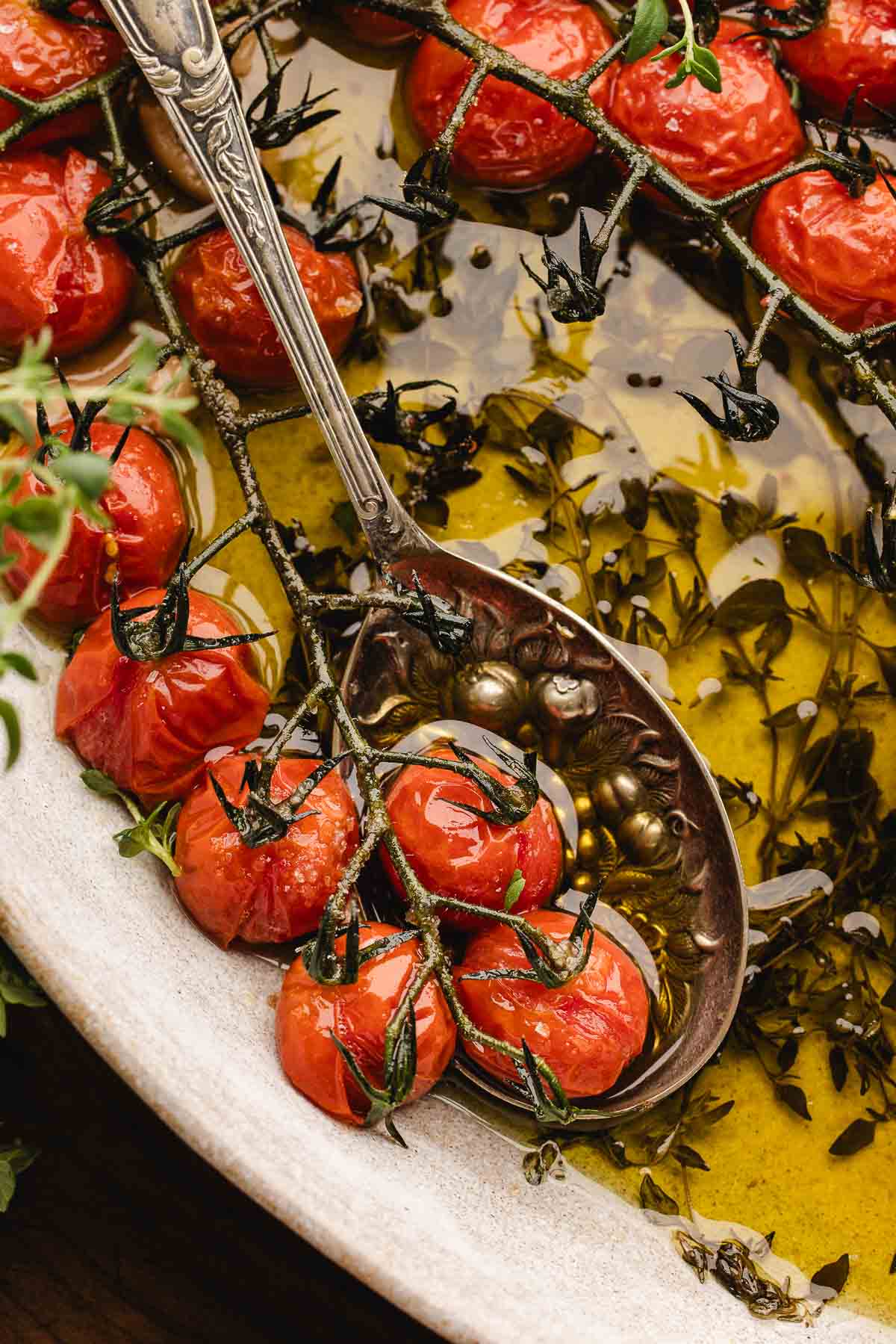 Roasted tomato confit sit in a dish of olive oil and herbs, with a metal spoon resting among them.