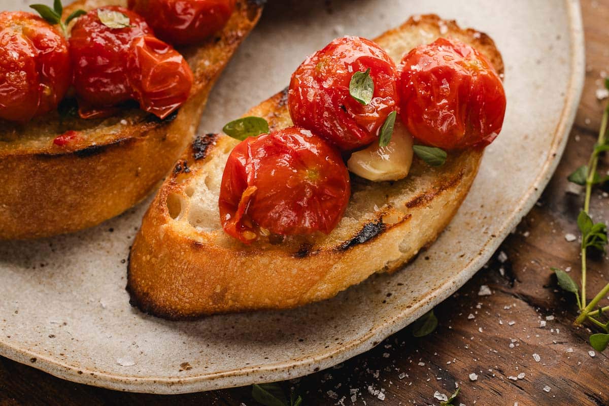 Toasted bread topped with tomato confit, and garlic is served on a ceramic plate, with scattered sea salt and herbs visible nearby.