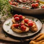 Three slices of toasted bread topped with tomato confit, with a bowl of tomatoes and fresh herbs in the background.