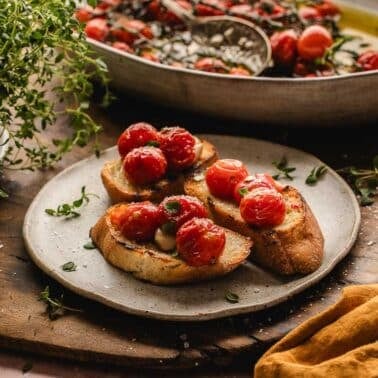 Three slices of toasted bread topped with tomato confit, with a bowl of tomatoes and fresh herbs in the background.