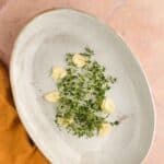 Oval ceramic plate with sprigs of fresh thyme and several peeled garlic cloves, placed on a pink-tiled surface with a mustard-colored cloth nearby.