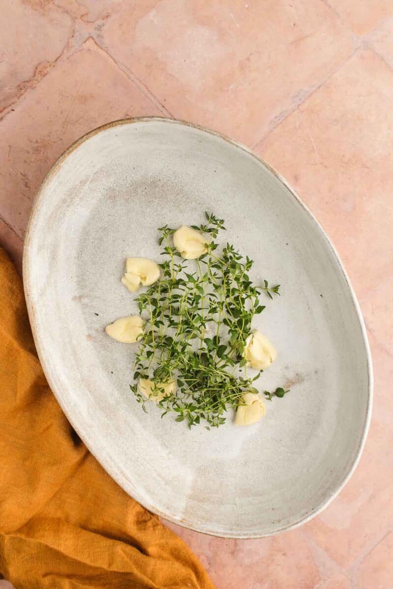 Oval ceramic plate with sprigs of fresh thyme and several peeled garlic cloves, placed on a pink-tiled surface with a mustard-colored cloth nearby.