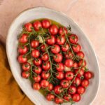 Oval dish filled with vine-ripened cherry tomatoes, drizzled with olive oil, placed on a light pink tiled surface with a mustard yellow cloth nearby.