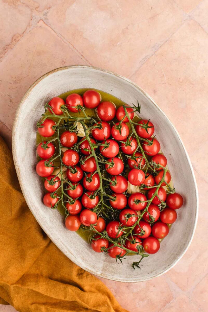 Oval dish filled with vine-ripened cherry tomatoes, drizzled with olive oil, placed on a light pink tiled surface with a mustard yellow cloth nearby.