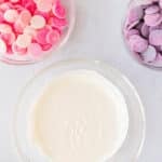 Four bowls with ingredients: pink and white melting wafers, purple melting wafers, white melted chocolate, and colorful sprinkles, on a light surface.