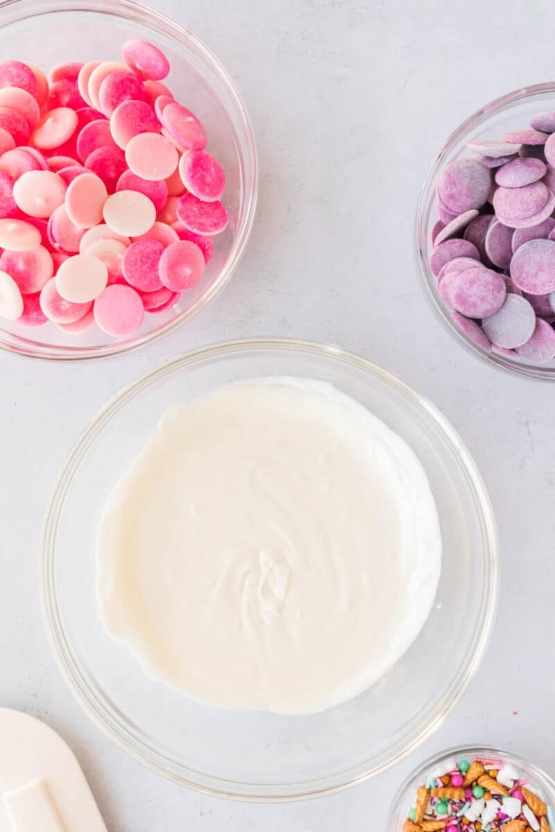 Four bowls with ingredients: pink and white melting wafers, purple melting wafers, white melted chocolate, and colorful sprinkles, on a light surface.