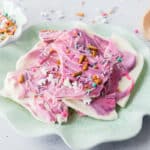 A scalloped plate holds pieces of Unicorn bark topped with colorful sprinkles and pretzel bits. A bowl of extra sprinkles and a scoop are in the background.