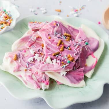 A scalloped plate holds pieces of Unicorn bark topped with colorful sprinkles and pretzel bits. A bowl of extra sprinkles and a scoop are in the background.