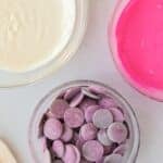 Three bowls: one with melted white chocolate, one with melted pink candy coating, and one with purple candy melts, all on a light countertop.