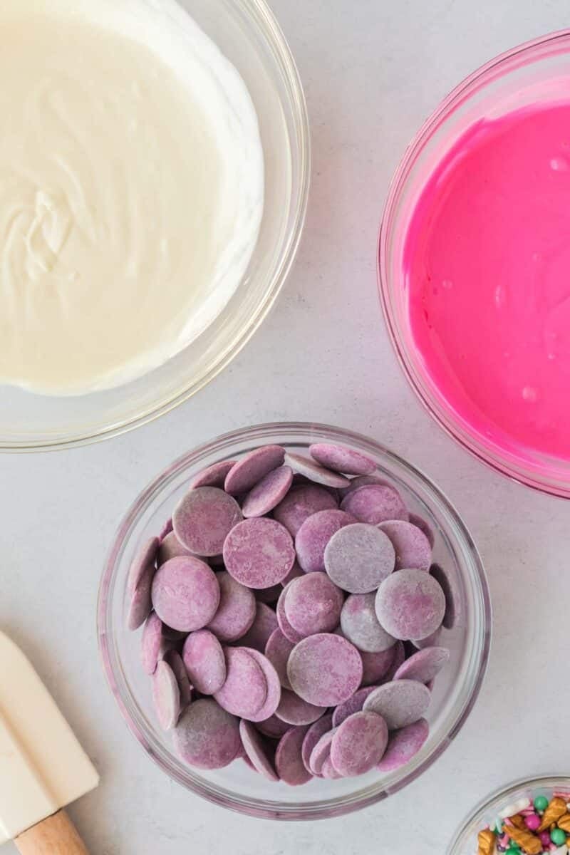 Three bowls: one with melted white chocolate, one with melted pink candy coating, and one with purple candy melts, all on a light countertop.