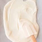 A white spatula spreads melted white chocolate on a sheet of parchment paper.