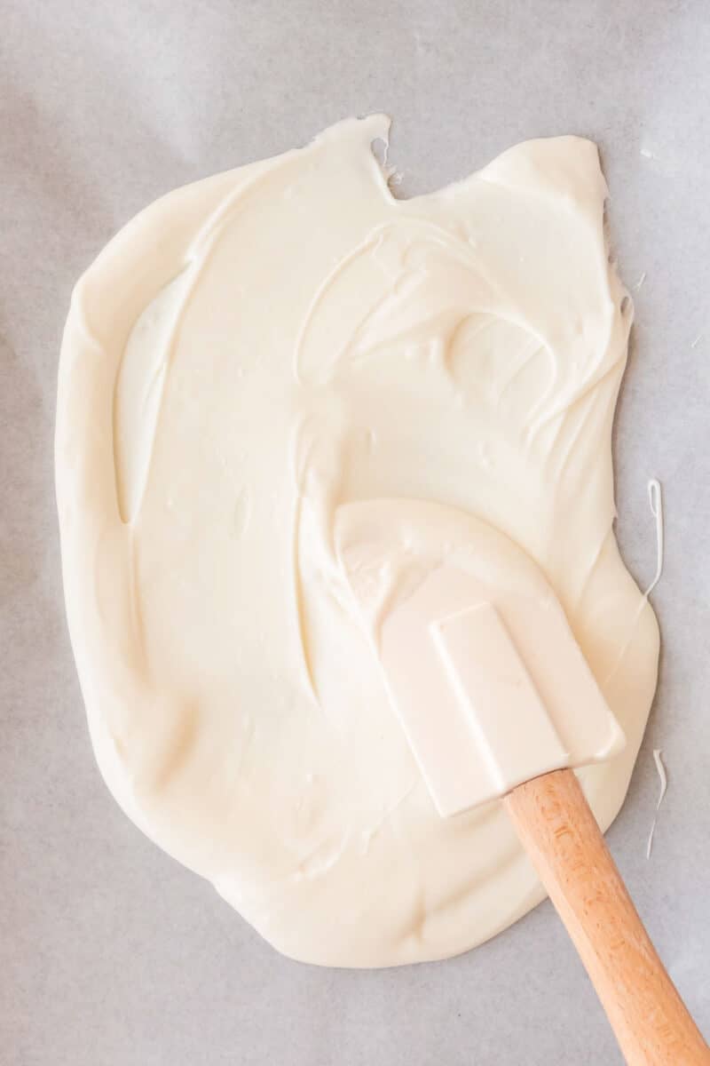 A white spatula spreads melted white chocolate on a sheet of parchment paper.