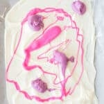 White melted chocolate spread on parchment paper with blobs and swirls of pink and purple melted chocolate on top.