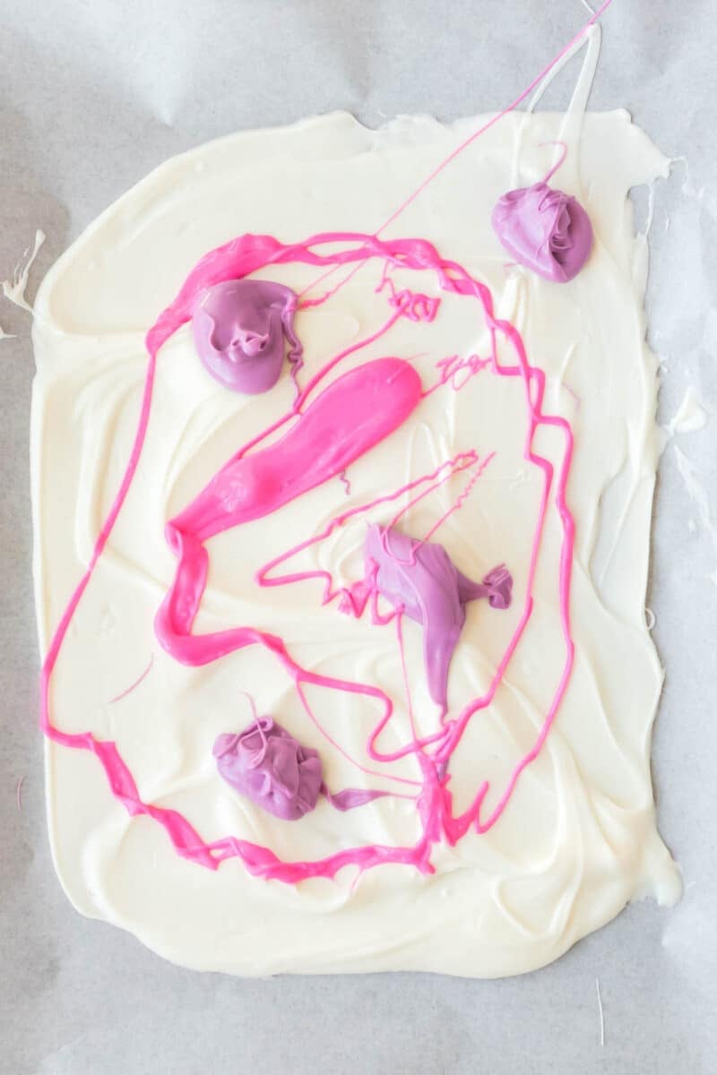 White melted chocolate spread on parchment paper with blobs and swirls of pink and purple melted chocolate on top.