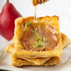 Square puff pastry tart topped with thin pear slices and herbs, stacked on a plate, with honey being drizzled over the top.