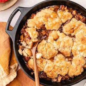 A cast iron skillet filled with baked apple cobbler, featuring golden biscuit topping and caramelized apple filling, with a wooden spoon resting inside.