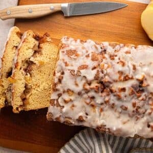 A glazed loaf cake with apple chunks, partially sliced on a wooden cutting board, with a knife and a striped cloth nearby.