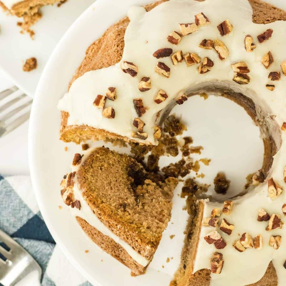 A bundt cake with white icing and chopped pecans on top, served on a white plate with a slice cut out.