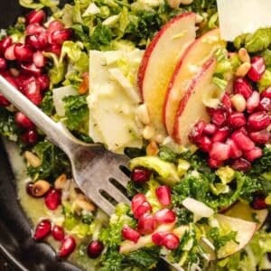 Salad with kale, pomegranate seeds, apple slices, pine nuts, and shaved cheese, with a fork and creamy dressing.