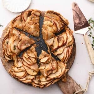A sliced caramel apple tart on a round wooden cutting board, with a pie server and sprigs of herbs nearby.