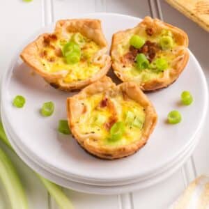 Three mini quiches topped with chopped green onions are arranged on a white plate, sitting on a white surface with additional green onions nearby.