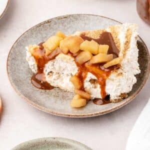 A slice of cheesecake topped with diced apples and caramel sauce sits on a ceramic plate.