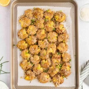 Smashed potatoes topped with grated cheese and chopped chives are arranged on a parchment-lined baking tray.