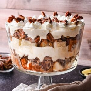 A glass trifle bowl filled with layers of cake, cream, chopped pecans, and whipped topping, set on a dark surface with extra pecans in a small bowl nearby.