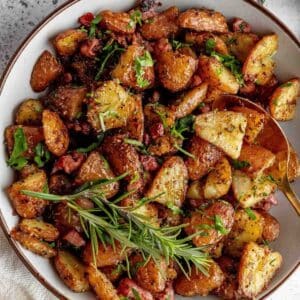 A bowl of roasted potatoes mixed with herbs, diced meat, and garnished with fresh parsley and a sprig of rosemary.