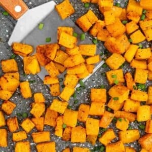 Roasted butternut squash cubes on a baking tray with chopped green herbs, and a metal spatula lifting some pieces.