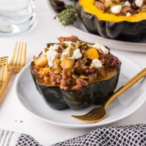 Half of a baked acorn squash stuffed with sausage, apples, and vegetables, topped with crumbled cheese and seeds, served on a white plate with a gold fork.