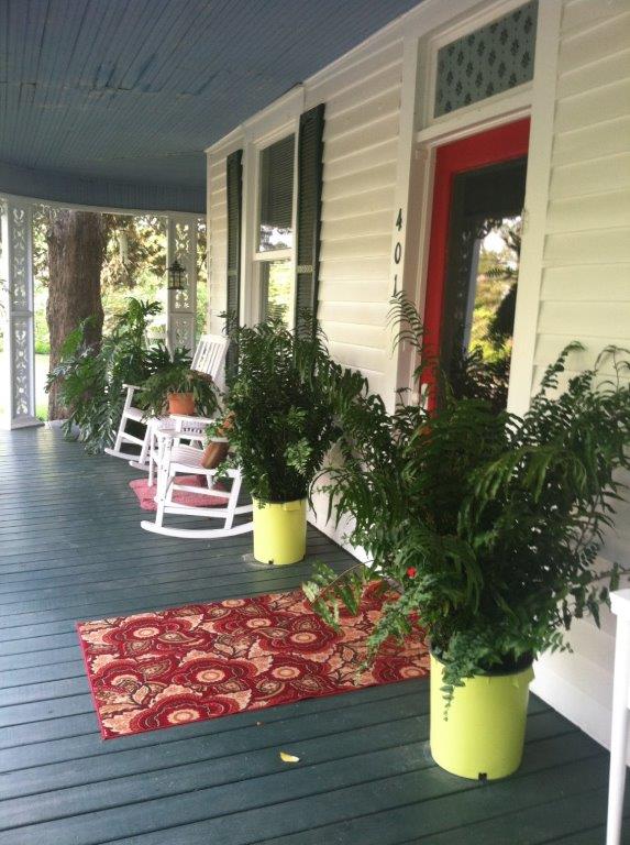 The front porch of a white farmhouse with rocking chairs, hanging ferns, an American flag, and pots of mums lining the steps.