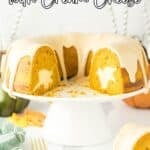 Pumpkin bundt cake with cream cheese filling and glaze sits on a white cake stand with a slice cut out, surrounded by autumn decor.