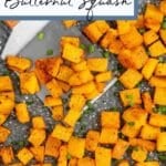 Diced roasted butternut squash pieces on a speckled baking sheet, garnished with chopped green herbs.