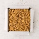 A square baking pan lined with parchment paper contains an unbaked crumble mixture spread evenly on top.