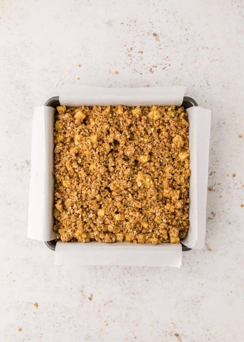 A square baking pan lined with parchment paper contains an unbaked crumble mixture spread evenly on top.