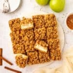 A plate of apple cheesecake bar, some cut into squares, surrounded by green apples, cinnamon sticks, and stacked plates with forks on a light surface.