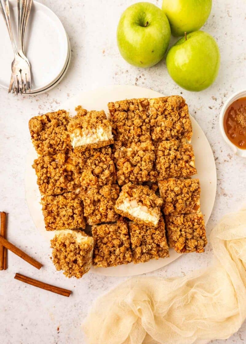 A plate of apple cheesecake bar, some cut into squares, surrounded by green apples, cinnamon sticks, and stacked plates with forks on a light surface.