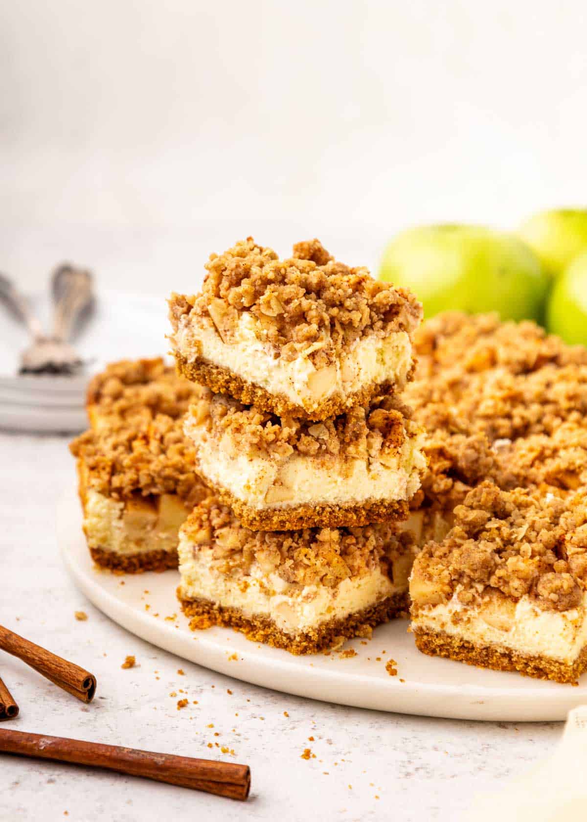 Three apple crumble cheesecake bars are stacked on a plate with more bars, green apples, cinnamon sticks, and forks in the background.
