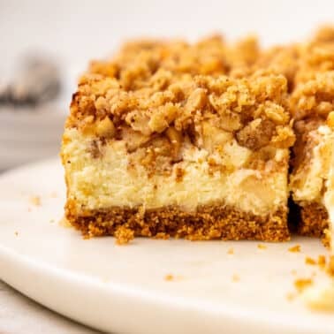 Close-up of an apple cheesecake bar with a creamy filling, a streusel topping, and a graham cracker crust on a white plate.