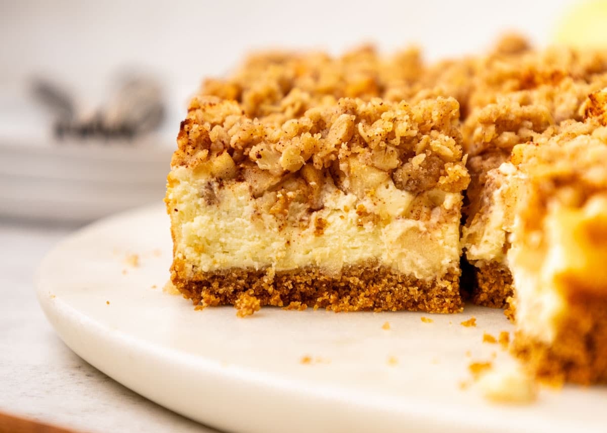 Close-up of an apple cheesecake bar with a creamy filling, a streusel topping, and a graham cracker crust on a white plate.