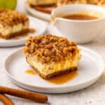 A square apple cheesecake bar with crumb topping and caramel sauce on a white plate, with cinnamon sticks and more dessert in the background.