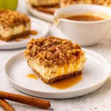 A square apple cheesecake bar with crumb topping and caramel sauce on a white plate, with cinnamon sticks and more dessert in the background.