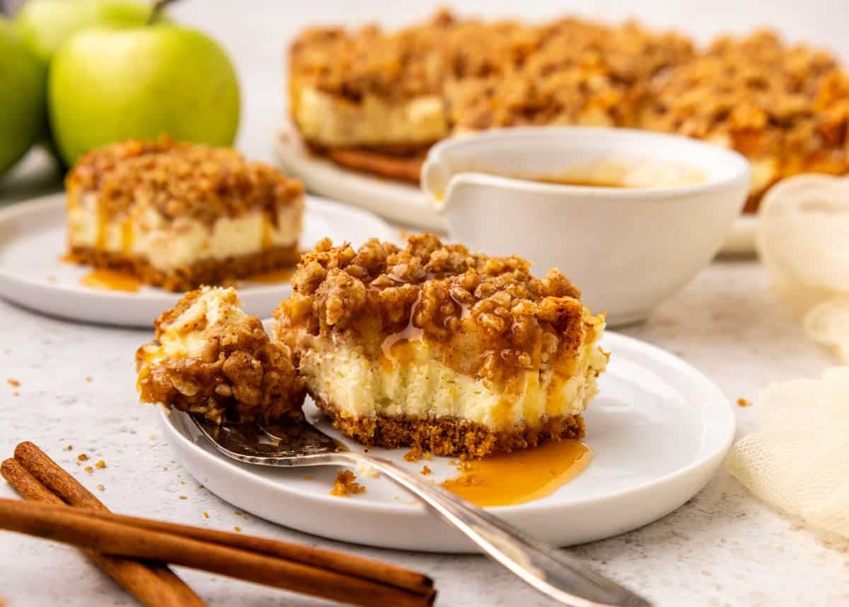 A slice of apple cheesecake bar with caramel sauce on a white plate, next to a fork, with apples and cinnamon sticks nearby.