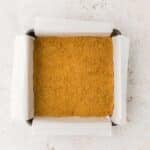 Square baking pan lined with parchment paper containing an even layer of finely crushed graham cracker crust on a light surface.