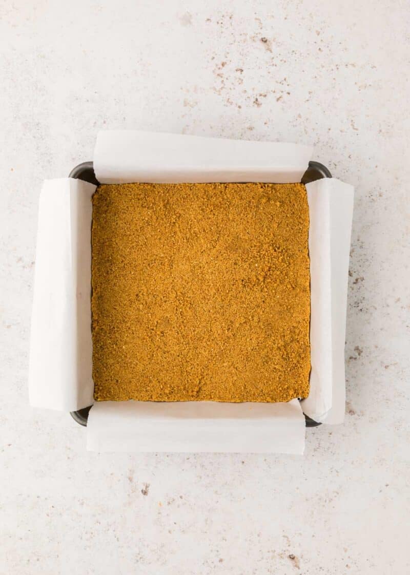 Square baking pan lined with parchment paper containing an even layer of finely crushed graham cracker crust on a light surface.