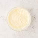 A glass bowl filled with whipped cream or frosting sits on a light, textured surface.