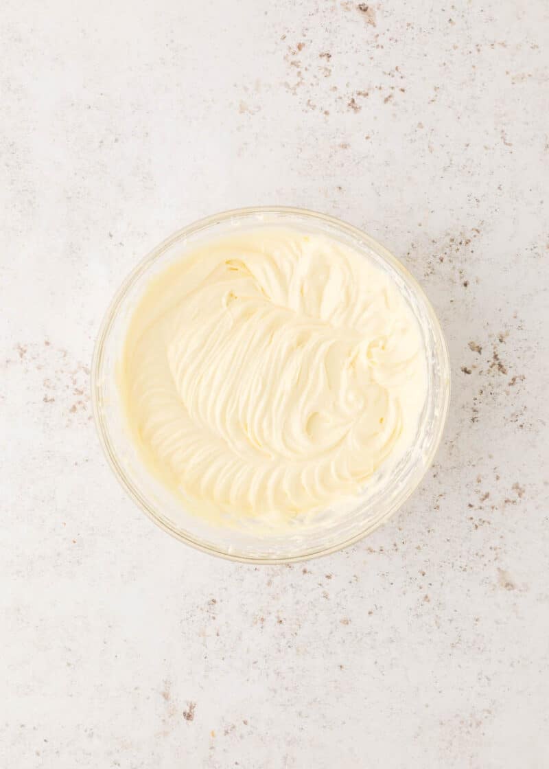 A glass bowl filled with whipped cream or frosting sits on a light, textured surface.