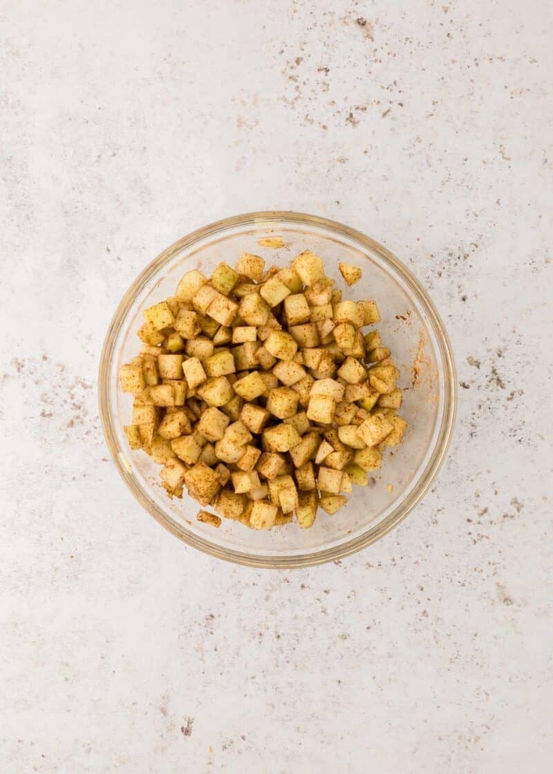 A glass bowl filled with small, diced apple pieces coated in brown spices on a light-colored surface.